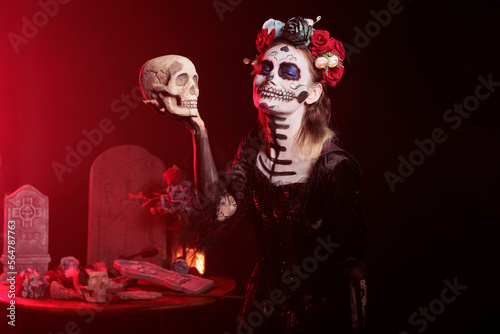 Creepy young person holding skull in studio, acting scary and horror to celebrate mexican halloween. Beautiful woman in festival costume with body art, looking like goddess of death.