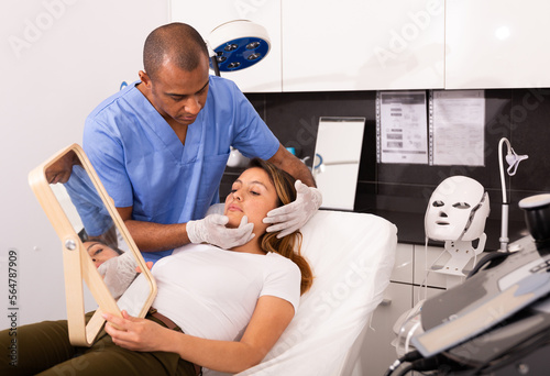 Confident cosmetologist examining female client face before procedure in esthetic clinic. Woman looking in mirror