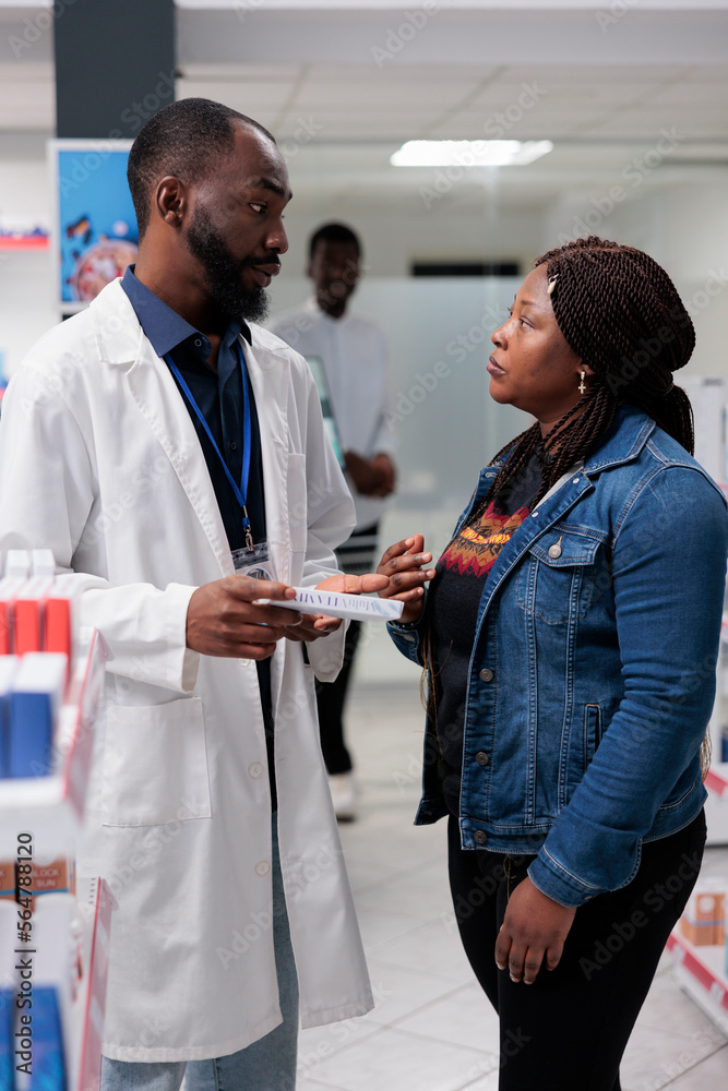 Fototapeta premium Pharmacy customer buying medication, pharmacy consultant recommending buyer pills, side view. African american woman talking with man pharmacist in drugstore aisle, all black team