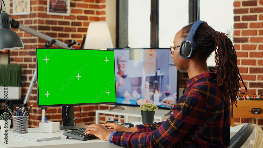 Female freelancer wearing headphones and using greenscreen on computer desktop, sitting at desk and working with isolated chroma key display. Looking at monitor with blank mock up.