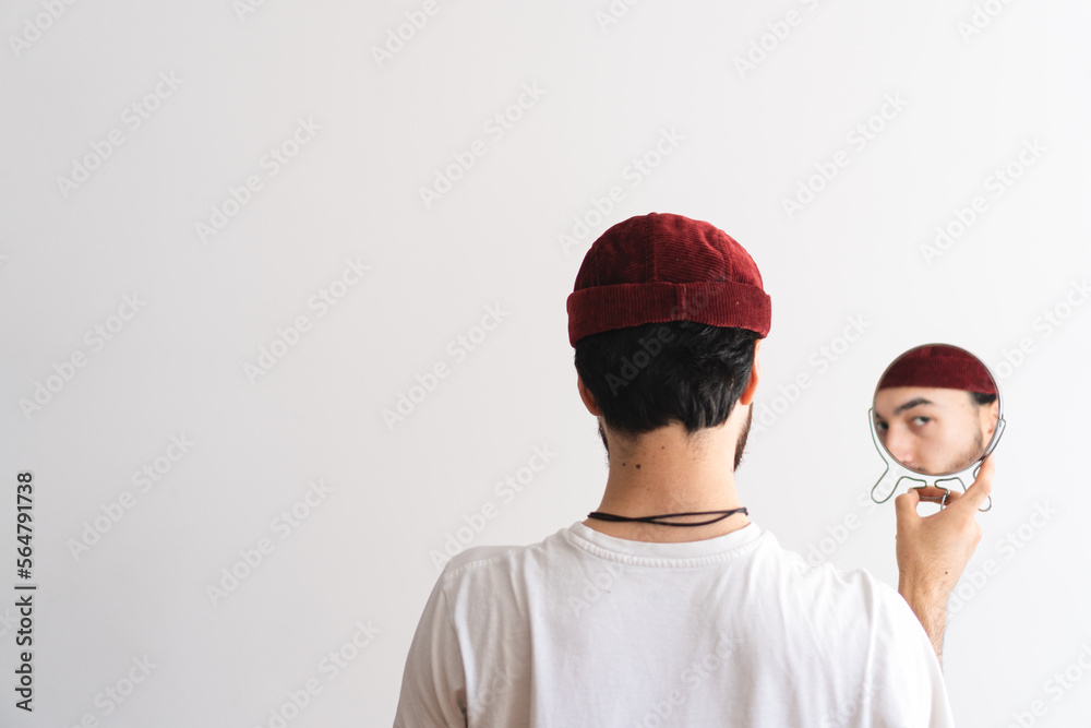 Young man with a raised eyebrow with his back turned wearing a brimless cap looking at camera through a mirror with a white background. Artistic photo. Copy space. Young man with a raised eyebrow with his back turned wearing a brimless cap looking at camera through a mirror with a white background. Artistic photo. Copy space.