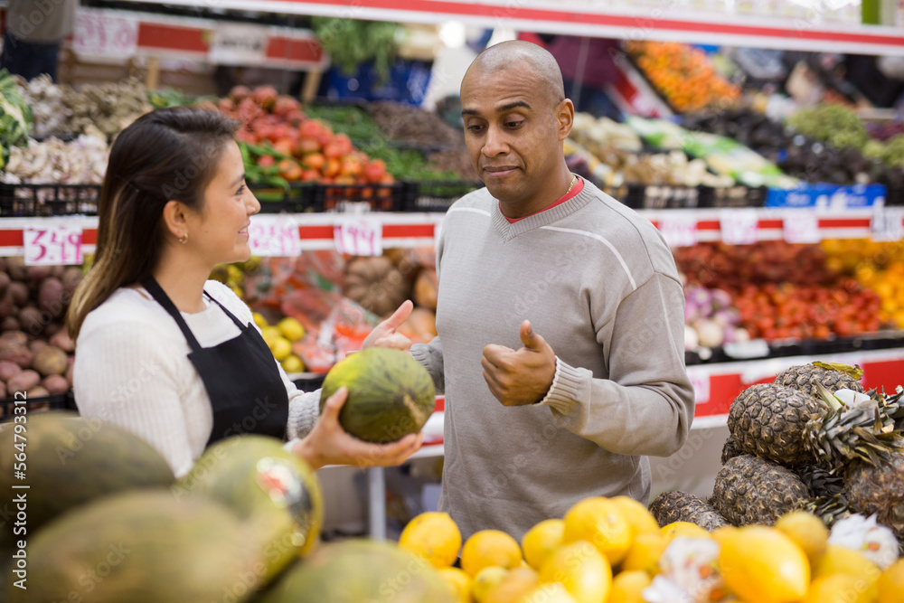 Latin man choosing ripe melons in supermarket and female store employee helping him