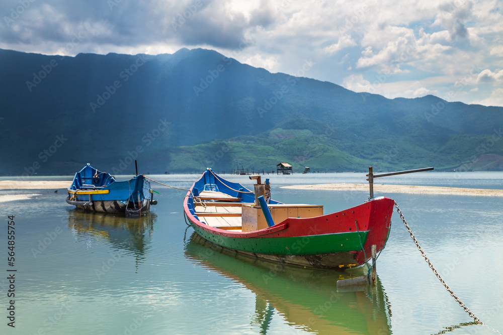 Two colourful wooden fishing boats on a calm coastal lagoon with rays of light breaking through clouds over distant mountains