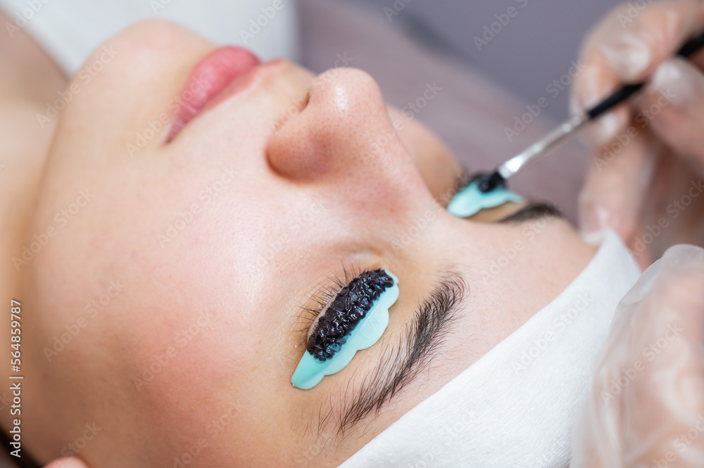 Close-up portrait of a woman on eyelash lamination procedure. The master applies tint to the eyelashes. 