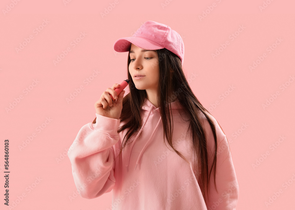 Young woman in cap with disposable electronic cigarette on pink background Young woman in cap with disposable electronic cigarette on pink background