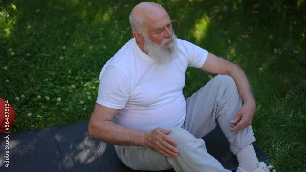 High angle view of old man stroking injured knee sitting on exercise mat on green summer meadow. Portrait of senior Caucasian sportsman with beard and grey hair caressing painful joint in slow motion
