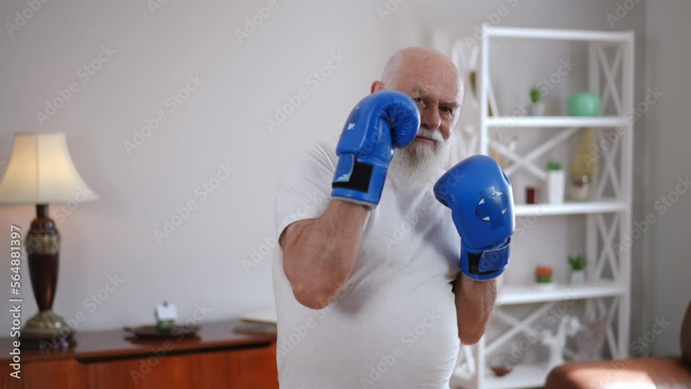 Bearded grey-haired Caucasian man posing in boxing gloves indoors smiling. Portrait of inspired motivated senior boxer looking at camera gesturing in slow motion. Martial arts and aging Bearded grey-haired Caucasian man posing in boxing gloves indoors smiling. Portrait of inspired motivated senior boxer looking at camera gesturing in slow motion. Martial arts and aging