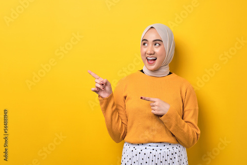 Beautiful shocked Asian girl wearing hijab pointing finger at empty copy space isolated on yellow background photo