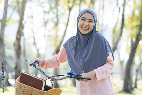 Smiling Asian young Muslim woman wear hijab headscarf and hijab dress walking with her bicycle and books in the park