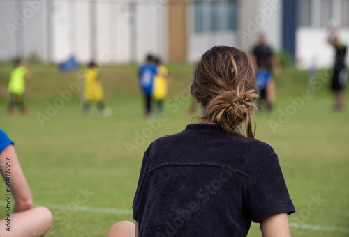 Mom and dad sitting and watching their son playing football in a school tournament on a sideline with a sunny day. Sport, outdoor active, lifestyle, happy family and soccer mom and soccer dad concept.