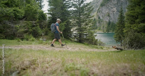 Hiker with the hat, backpack and hiking poles walking towards the mountain lake, surrounded by conifers, mountain the back.