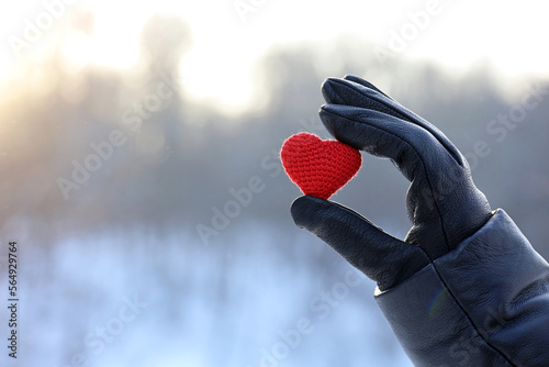 Red knitted heart in female hand in black leather glove on snow park and sun background. Concept of romantic love, Valentine's day, winter weather