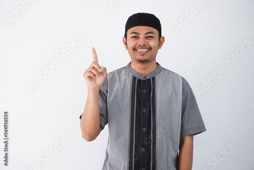 young asian muslim man finger pointing up raising a finger got a good idea looks surprised with a smile wearing grey koko clothes, isolated on a white background. photo