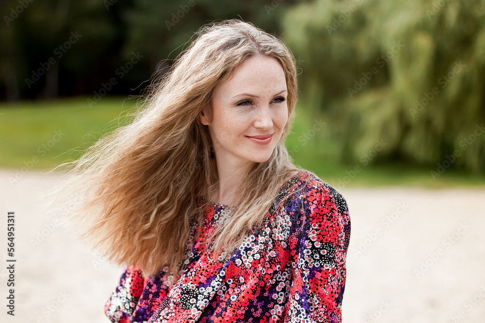Fototapeta premium Portrait of a cute smiling girl with long blond hair blowing in the wind, in a red summer sundress on the beach, sand and trees all around.