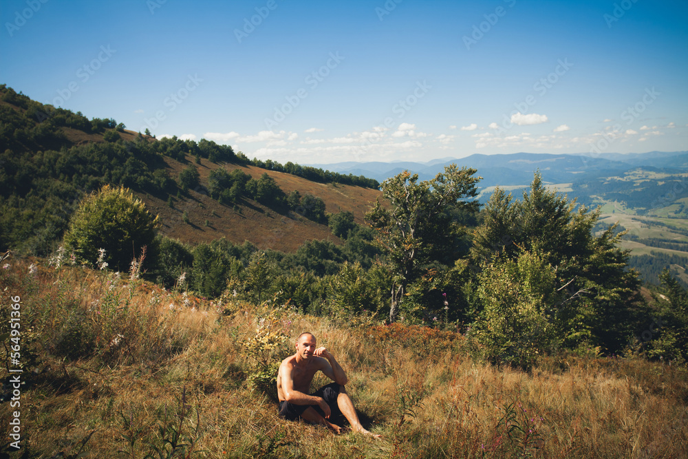 A man of athletic build in dark shorts sits on the grass in the mountains, hot summer. Around the trees and blue sky with clouds.