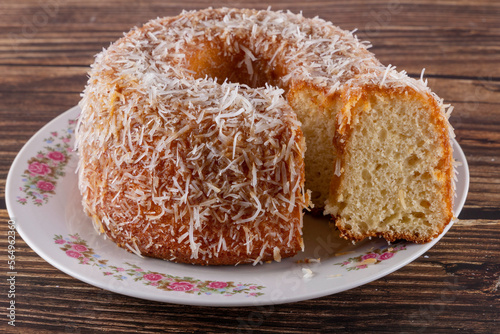 Coconut-flavored sweet cake topped with coconut flakes, with a cut slice showing the delicious dough on a decorated plate. Using wooden board and wooden background. Selective focus.