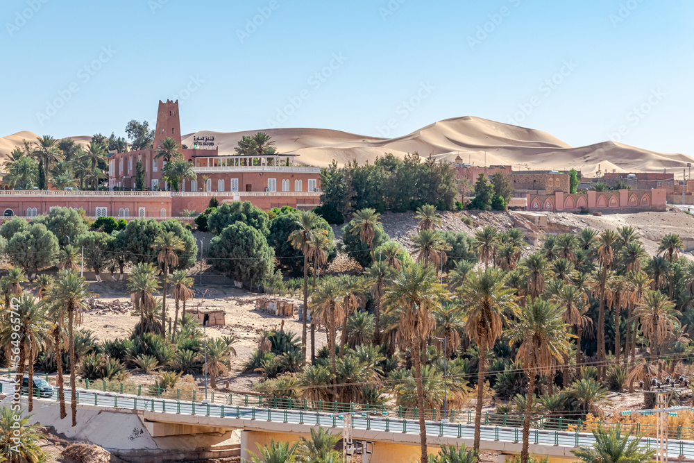 Taghit town of Bechar province in Algeria Sahara desert. Palm trees oasis, sand dunes and buildings. Road bridge of Oued Zouzfana river and Saoura hotel with an Algerian flag on the roof.