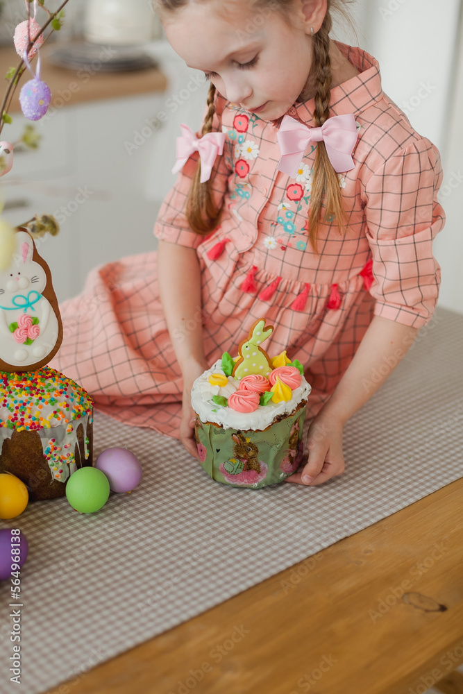 A young happy beautiful girl with 2 braids in peach colored dress is holding Easter cake. Close up. colorful eggs and tree branches for Easter home decoration. Kitchen in the background