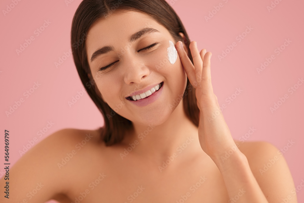 Delighted young woman applying cosmetics on face and smiling in pink studio Delighted young woman applying cosmetics on face and smiling in pink studio