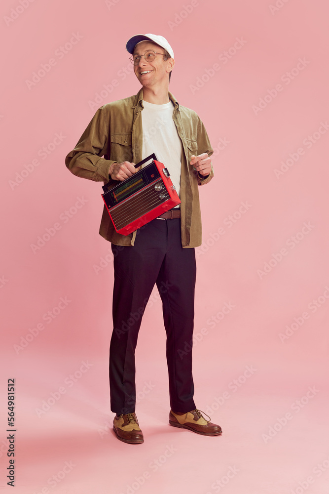 Cheerful mood. Young positive man in shirt, trousers and cap posing with vintage radio set over pink studio background. Music. Concept of emotions, facial expression, lifestyle, retro fashion. Ad Cheerful mood. Young positive man in shirt, trousers and cap posing with vintage radio set over pink studio background. Music. Concept of emotions, facial expression, lifestyle, retro fashion. Ad
