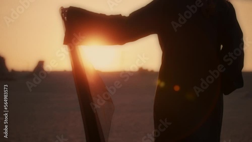 A girl holding a black scarf in her hand walking barefoot in the desert wearing an abaya while sunset stunning lens flare medium shot fossil dunes photo