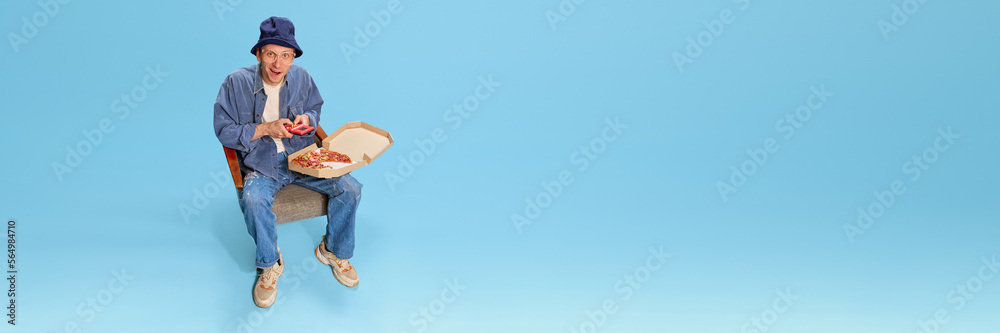 Cheerful man in jeans clothes sitting with pizza and playing retro game console over blue studio background. Concept of emotions, facial expression, lifestyle, retro fashion. Ad. Banner Cheerful man in jeans clothes sitting with pizza and playing retro game console over blue studio background. Concept of emotions, facial expression, lifestyle, retro fashion. Ad. Banner