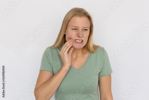 Frustrated young woman suffering from toothache. Portrait of sick Caucasian female model with fair hair in green T-shirt looking away, frowning and touching jaw. Despair, pain concept