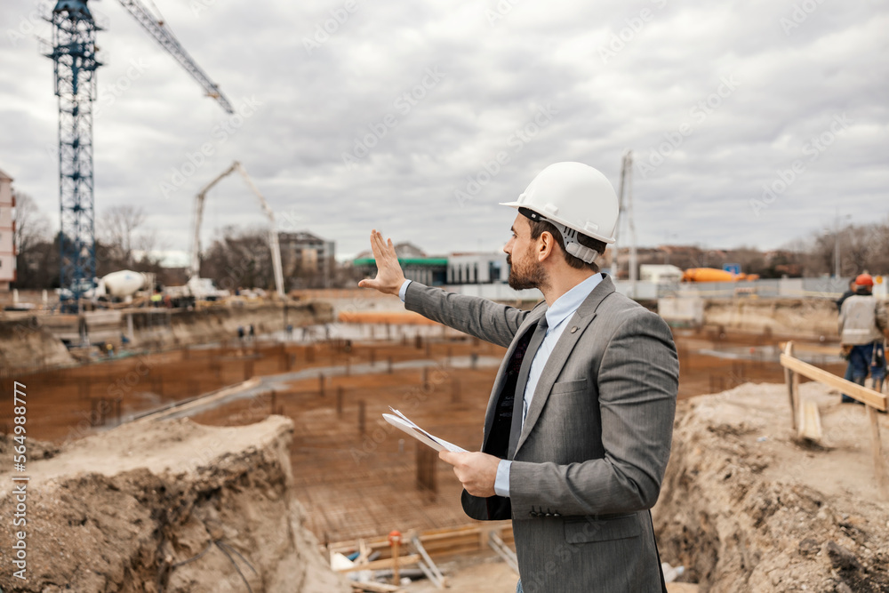 An architect is looking at the building in building process with documents in his hands.