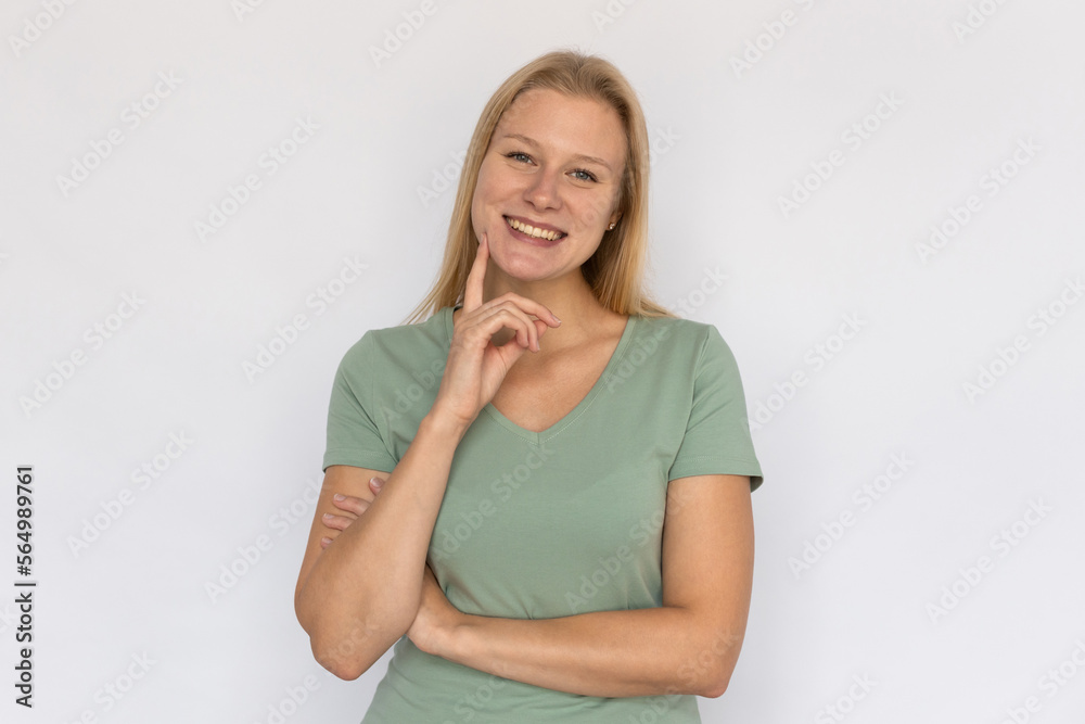Happy young woman with finger on chin. Portrait of pleased Caucasian female model with fair hair in green T-shirt looking at camera, smiling, touching face. Skincare, happiness concept Happy young woman with finger on chin. Portrait of pleased Caucasian female model with fair hair in green T-shirt looking at camera, smiling, touching face. Skincare, happiness concept