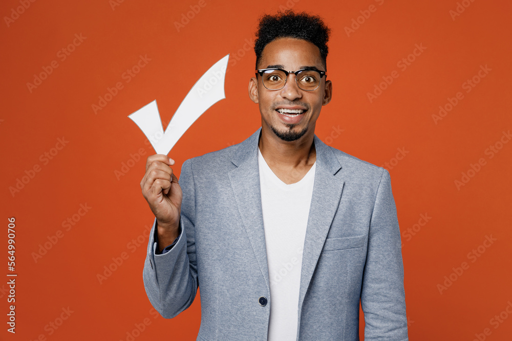 Young surprised fun employee business man corporate lawyer wear classic formal grey suit shirt glasses work in office hold in hand white check mark sign isolated on plain red orange background studio. Young surprised fun employee business man corporate lawyer wear classic formal grey suit shirt glasses work in office hold in hand white check mark sign isolated on plain red orange background studio.
