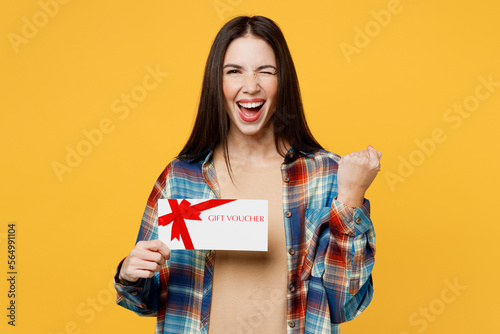 Young woman wear blue shirt beige t-shirt hold store gift certificate coupon voucher card do winner gesture wink blink eye isolated on plain yellow background studio portrait People lifestyle concept