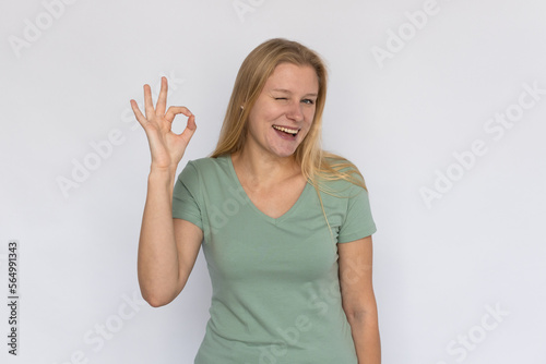 Excited young woman with ok gesture. Portrait of happy Caucasian female model with fair hair in green T-shirt looking at camera, smiling, winking with raised hand. Optimism, success concept