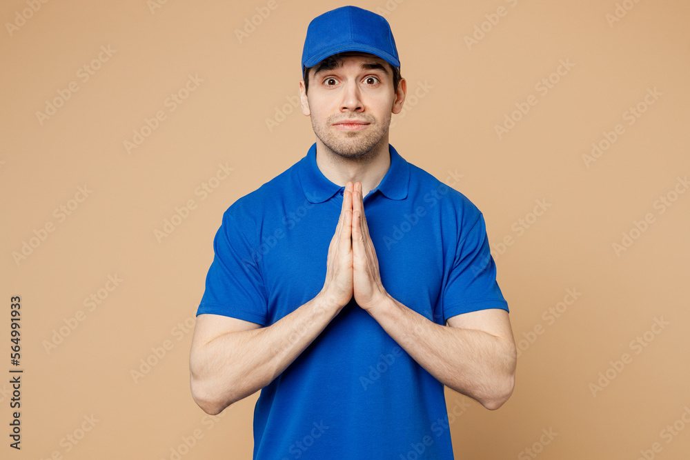 Delivery guy employee man wear blue cap t-shirt uniform workwear work as dealer courier hold hand folded in prayer gesture beg about something isolated on plain light beige background Service concept