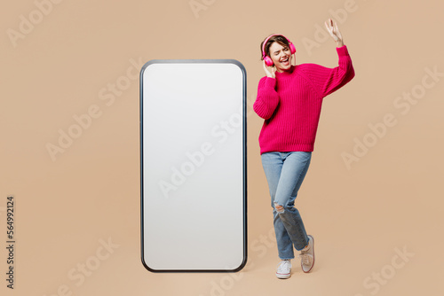 Full body young fun woman wear pink sweater headphones listen music big huge blank screen mobile cell phone smartphone with workspace copy space mockup area isolated on plain pastel beige background