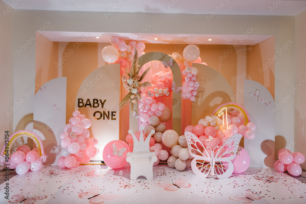 Foto Stock Children's photo zone. Birthday party for 1 year old girl on a  background photo wall. Arch decorated pink balloons, rainbow, text baby  one, flowers, paper decor butterfly, and wooden white