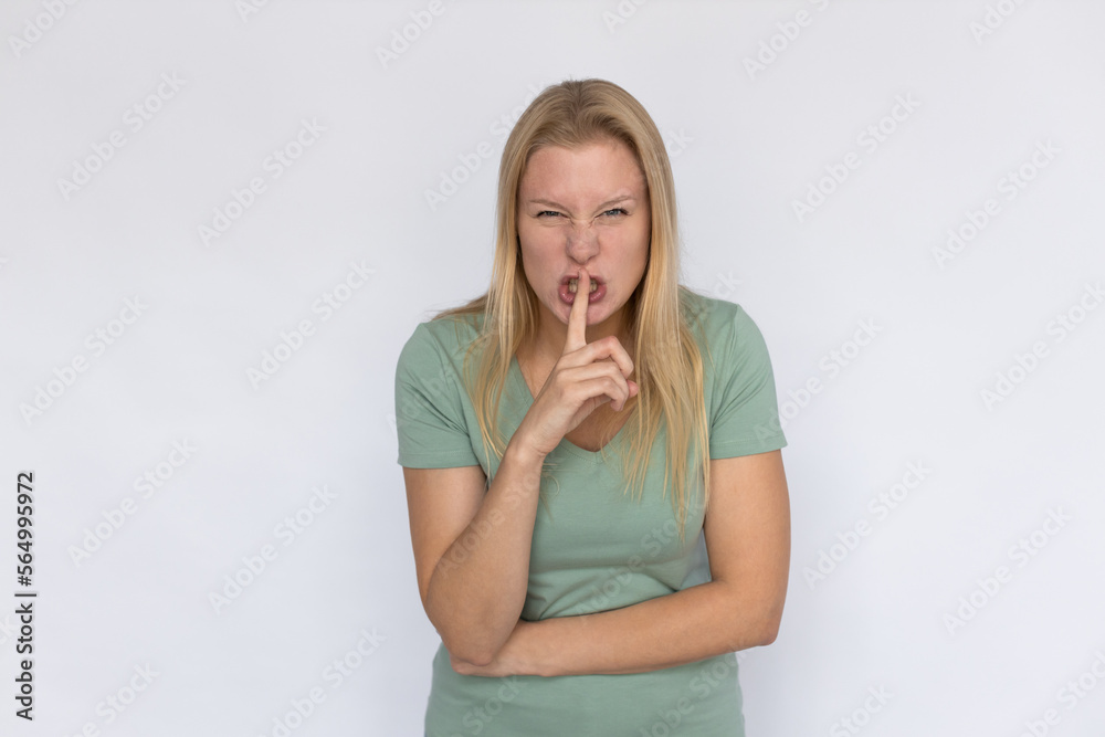 Angry young woman shushing. Portrait of displeased Caucasian female model with fair hair in green T-shirt looking at camera, pressing finger to lips and frowning. Silence, secret concept Angry young woman shushing. Portrait of displeased Caucasian female model with fair hair in green T-shirt looking at camera, pressing finger to lips and frowning. Silence, secret concept