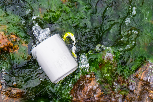 A Coffee mug laying out on the top of some rocks that covered with moss while the water keep splashing over it