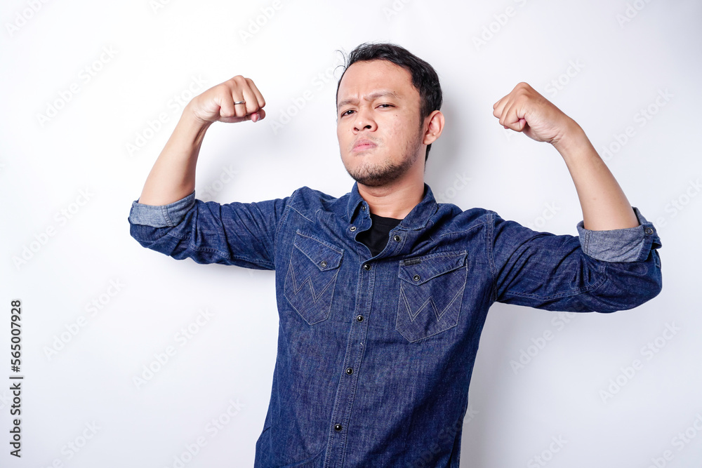 Fototapeta premium Excited Asian man wearing a blue shirt showing strong gesture by lifting his arms and muscles smiling proudly