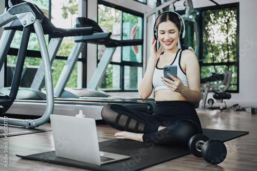 Happy young woman wearing wireless headset and listening to music while resting, sitting on fitness mat after exercising in gym indoors. © Thanumporn
