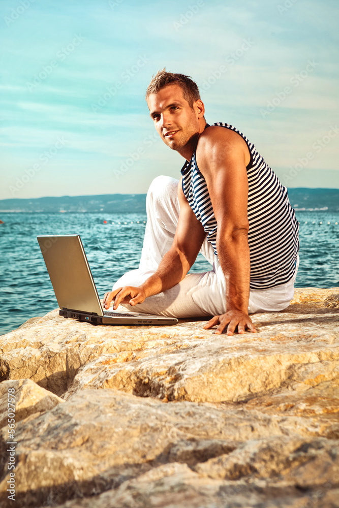 Man with the computer on the rocky beach wearing a sleeveless sailor shirt 