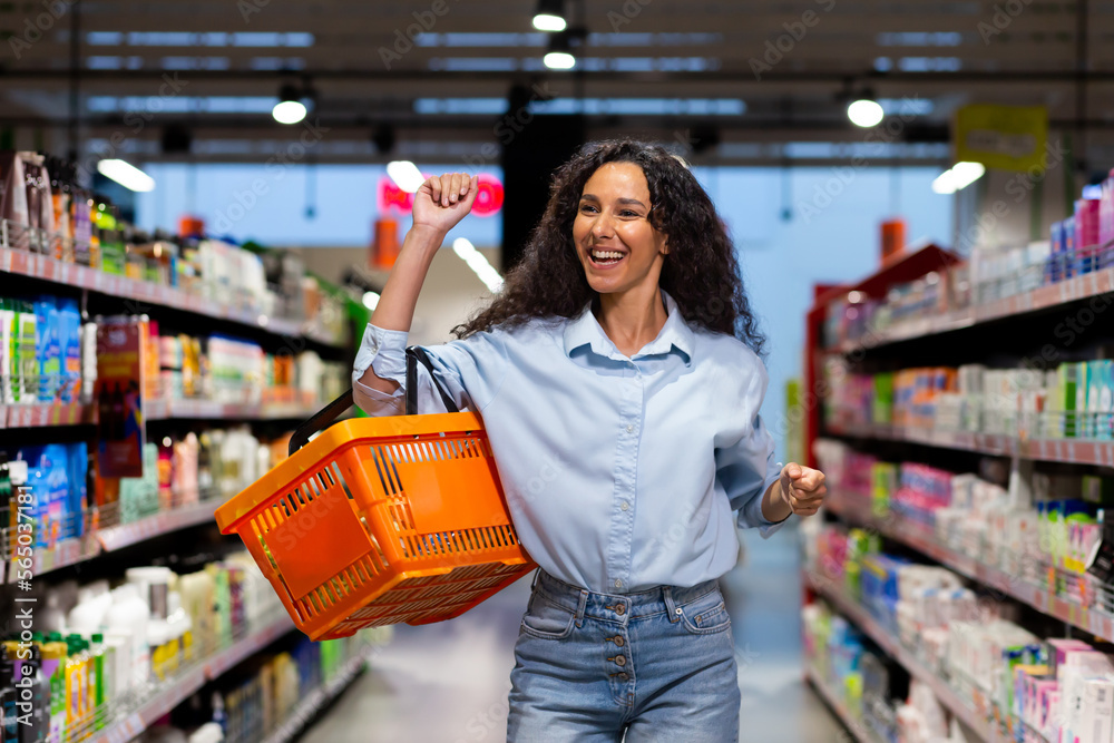Portrait of a happy woman shopper in a supermarket, a Hispanic woman with a basket of goods smiles with pleasure and dances among the shelves with goods, a satisfied shop customer.