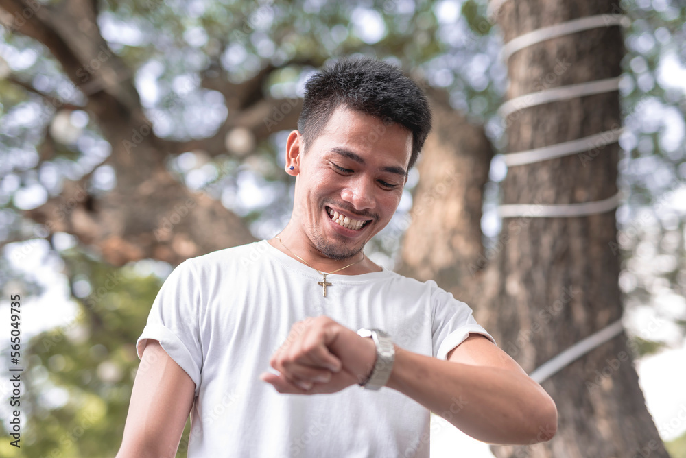 A smiling Southeast Asian man checks the time with his silver wristwatch in the middle of the day at the park to see if he is late for work. A smiling Southeast Asian man checks the time with his silver wristwatch in the middle of the day at the park to see if he is late for work.