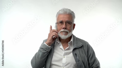 Portrait of an elderly retired man in glasses listens carefully on a mobile phone on a white background. Gray-haired grandfather with a beard posing on an isolated studio background. Slow motion. photo