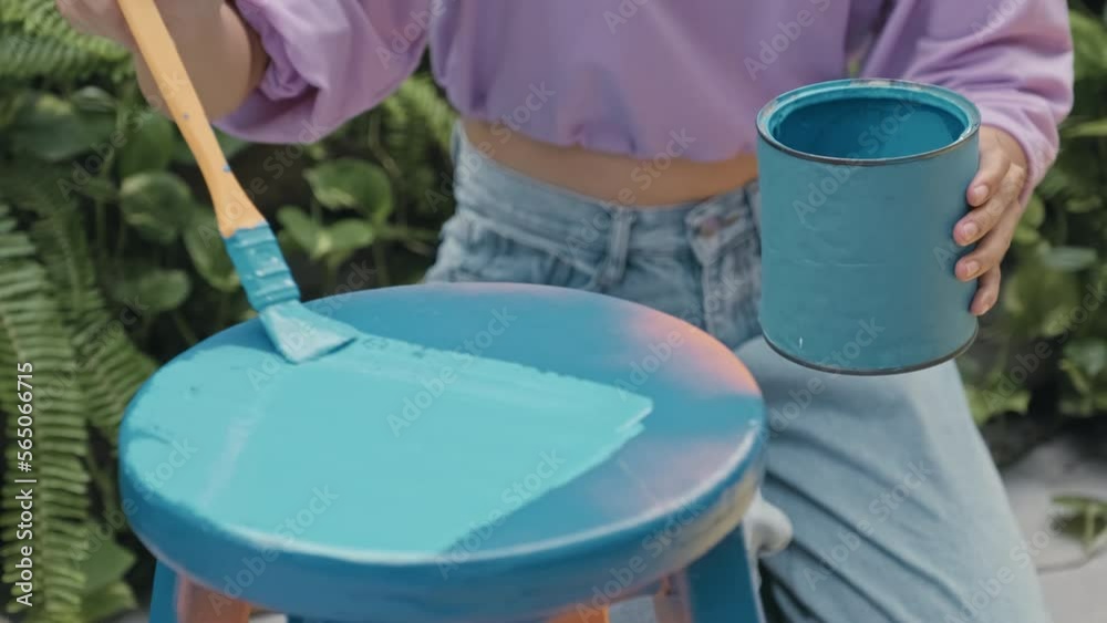 Cropped impersonal shot of girl painting old stool with blue paint while restoring furniture outdoors in backyard Cropped impersonal shot of girl painting old stool with blue paint while restoring furniture outdoors in backyard