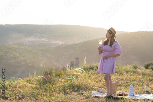 Woman in a straw hat and a purple dress stands in full growth and looks at the sunset with a glass of white wine. Summer holidays vacation relax in nature in the mountains. Concept: summer vacation 