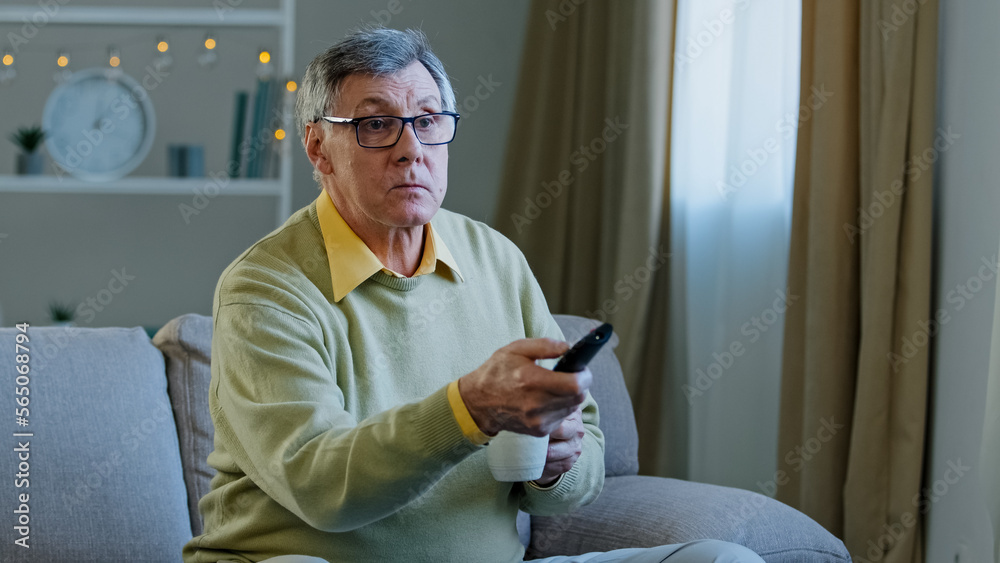 Caucasian aged senior mature man in glasses relax on couch at home change  channel with remote controller watching TV program relaxing with drinking  tea coffee old male choose movie television indoors Stock