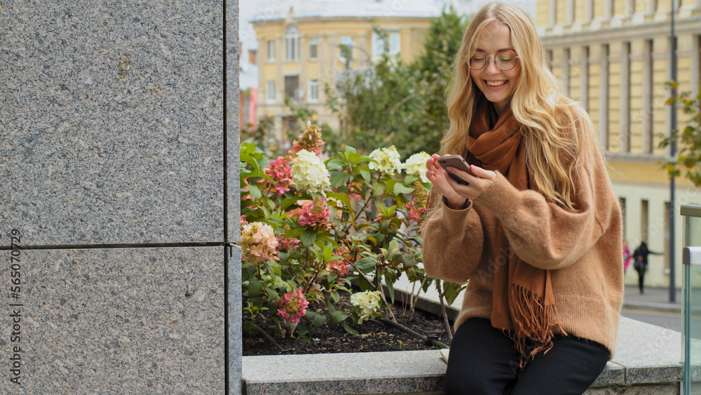 Fototapeta premium Caucasian woman 20s girl in glasses sitting outdoors in city browsing scrolling smartphone smiling female lady shopping online with phone mobile store app chatting with friend in cell social media