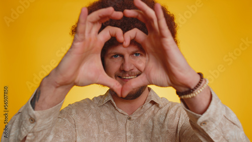 Man in love. Smiling man with lush afro hairstyle coiffure makes heart gesture demonstrates love sign expresses good feelings and sympathy. Young guy boy isolated alone on yellow studio background