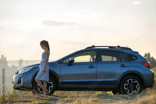 Happy young woman driver in blue dress enjoying warm summer evening standing beside her car. Travelling and vacation concept