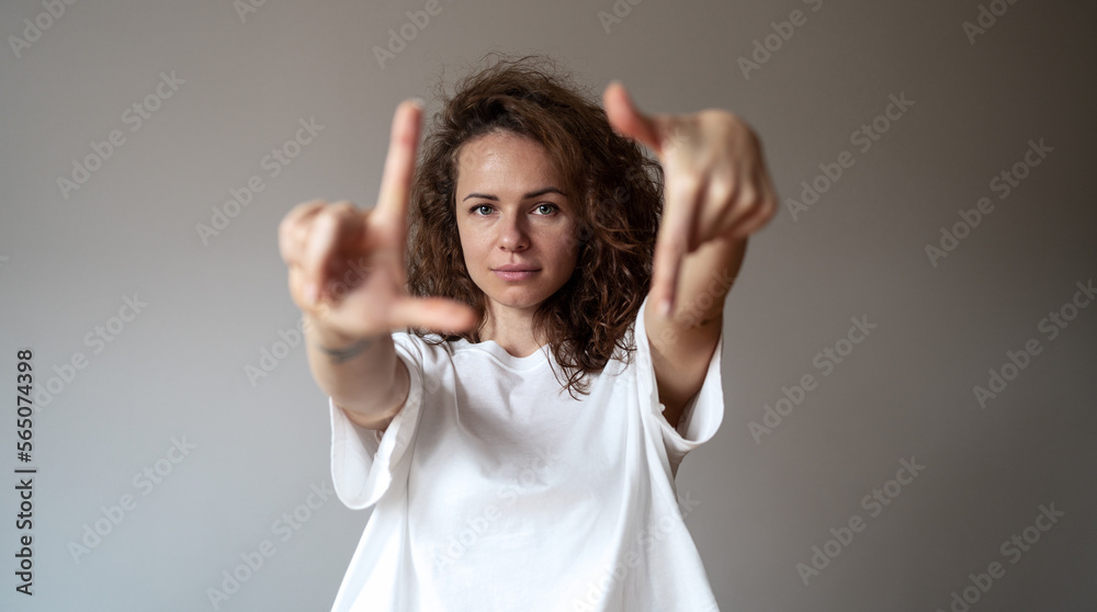 Young brunette curly hair woman wearing white tee shirt looking at camera through her fingers against plain grey background. Young brunette curly hair woman wearing white tee shirt looking at camera through her fingers against plain grey background.